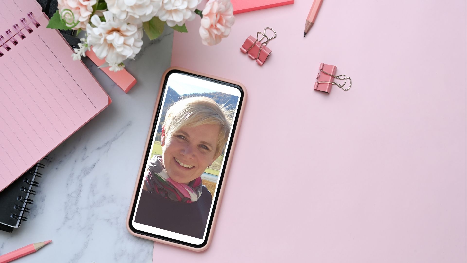 Tanja Scheicher in einem Handy umgeben von rosa Schreibtischutensilien wie Klammern, Bleistift Radiergummi und Notizblock.