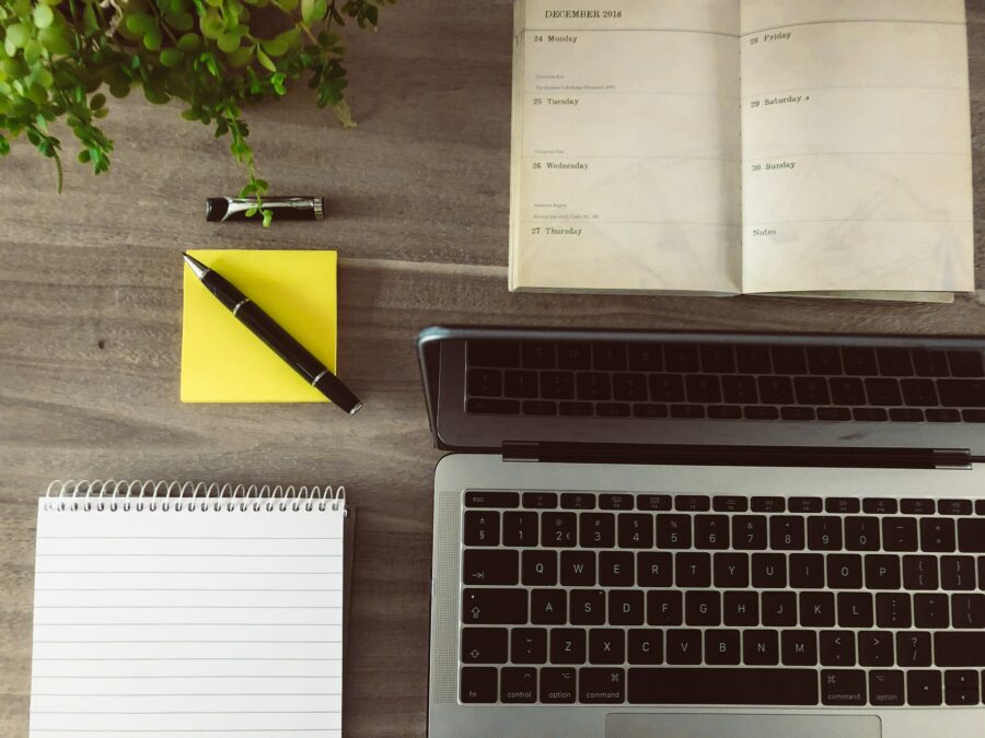 Es liegen ein Block, ein Kalender, ein Haftnotizblock mit einem Stift und ein Laptop auf einem Tisch.